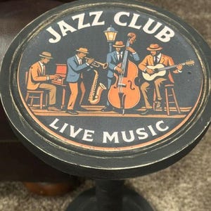 May include: A round black side table with a distressed finish featuring a graphic of a jazz band. The illustration shows four musicians playing a piano, saxophone, double bass, and guitar, with the text JAZZ CLUB and LIVE MUSIC printed.