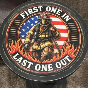 May include: A round black wooden sign featuring a graphic of a firefighter in tan gear standing in front of an American flag. The design includes orange flames at the bottom and the white text "FIRST ONE IN" at the top and "LAST ONE OUT" at the bottom.