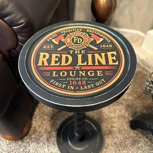 May include: A round black side table with a pedestal base. The tabletop features a graphic design with the text "THE RED LINE LOUNGE," "PRIDE TRADITION BROTHERHOOD," "EST. 1648," "ENGINE CO. 1648," and "FIRST IN LAST OUT" surrounding a central fire department emblem.