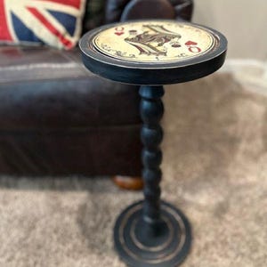 May include: A black pedestal side table with a round top featuring a Queen of Hearts playing card design. The table has a tall, slender, turned spindle-style leg and a circular base. The surface of the tabletop displays the classic red and black card illustration with ornate decorative scrollwork details.