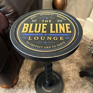 May include: A round black side table with a pedestal base. The tabletop features a circular graphic with the text "THE BLUE LINE LOUNGE" in gold, "DUTY INTEGRITY RESPECT" and "TO PROTECT AND TO SERVE" in white, and a blue horizontal line.