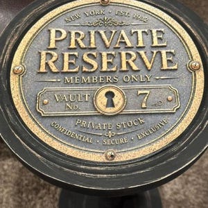 May include: A round black and gold decorative sign featuring the text NEW YORK EST. 1932 PRIVATE RESERVE MEMBERS ONLY VAULT No. 7 PRIVATE STOCK CONFIDENTIAL SECURE EXCLUSIVE with a central keyhole graphic and four visible metal screws on the border.
