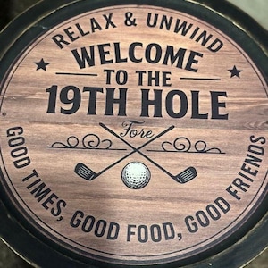 May include: Round wooden sign with the text "RELAX & UNWIND WELCOME TO THE 19TH HOLE" in black lettering. The sign features crossed golf clubs, a golf ball, and the words "GOOD TIMES, GOOD FOOD, GOOD FRIENDS".