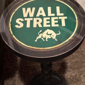 May include: A decorative table with a round top featuring the words "WALL STREET" in white letters on a green background. A white bull graphic is below the text. The table has a dark, ornate base.