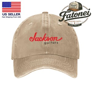Puede incluir: Gorra de b&eacute;isbol beige con la inscripci&oacute;n "Jackson guitars" en rojo. La gorra tiene una visera curva y un aspecto desgastado. El logotipo "Fatonei Custom Hats" es visible en la esquina superior derecha.