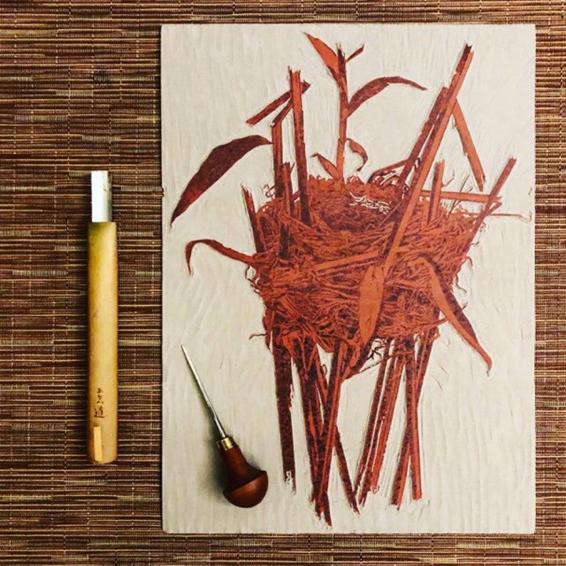 May include: A close-up of a woodblock print of a bird's nest made with twigs and leaves. The print is in shades of red and brown on a white background. Two carving tools are visible in the foreground.