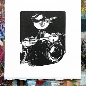 May include: Black and white linocut print of a small bird perched on top of a vintage Minolta camera. The bird is facing the left side of the image and the camera is facing the right side of the image.