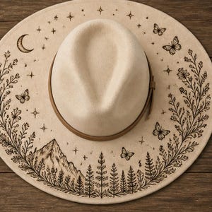 May include: A cream-coloured fedora hat with a brown leather band. The wide brim features a detailed black illustration of a mountain range, trees, butterflies, stars, and a crescent moon. The hat is set against a wooden background.
