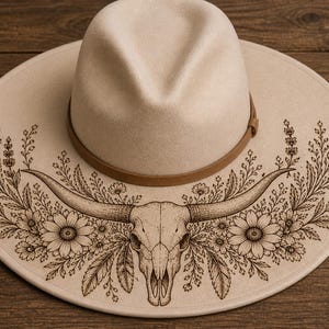 May include: A cream-colored fedora hat with a brown leather band. The hat's brim features a detailed laser-etched design of a bull skull, flowers, and foliage. The hat is a stylish accessory.