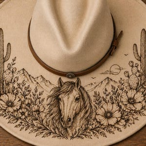 May include: A beige cowboy hat with a brown leather band. The hat features a detailed desert landscape design, including a horse, cacti, mountains, and flowers. The design is etched in a dark brown color.