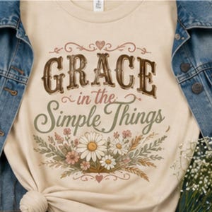 May include: A cream-colored t-shirt featuring the brown and green text "GRACE in the Simple Things" centered above a colorful illustration of daisies and wildflowers, layered underneath a blue denim jacket with metal buttons and visible stitching.