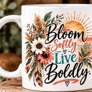 May include: A white ceramic mug featuring a colorful graphic of a sun, flowers, and feathers. The text reads Bloom Softly. Live Boldly. The design includes orange, red, green, and brown tones with a small heart icon below the text.