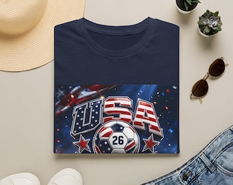May include: A navy blue t-shirt featuring a graphic print of a football with the number 26, surrounded by the text USA in a red, white, and blue flag pattern, accented with stars and confetti against a dark blue background with light flares.