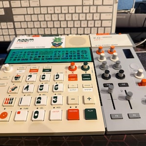 Könnte beinhalten: Ein weiß-graues elektronisches Musikproduktionsgerät mit Tastatur und Mixer. Das Gerät hat Knöpfe, Regler und Schieberegler in verschiedenen Farben, darunter Weiß, Grün, Orange und Grau. Der Text "RIDDIM" ist sichtbar.