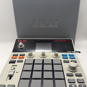 May include: A gray Akai MPC Sample music production center with a matching gray case. The MPC features numerous buttons, knobs, and pads in various colors. The text "AKAI" is visible on both the MPC and the case.