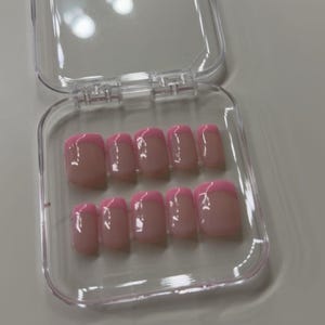 May include: A set of ten artificial nails in a clear plastic case. The nails are a light pink with a bright pink French tip. The nails are arranged in two rows of five.