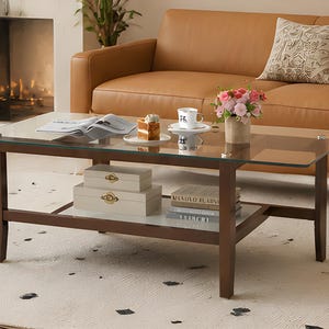 May include: A rectangular coffee table with a glass top and a lower shelf. The frame is dark brown. The table holds decorative boxes, books, a cup and saucer, a cake, and a vase of flowers. A tan sofa and patterned pillow are in the background.