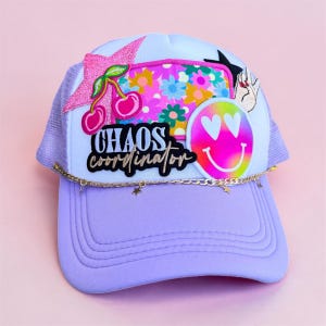 May include: A lilac trucker hat with a mesh back and a curved brim. The hat is decorated with patches including cherries, a floral design, a smiley face with heart eyes, and the text "CHAOS COORDINATOR". A gold chain with star charms adorns the brim.