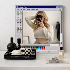 Op de afbeelding: Decoratieve spiegel ontworpen als een vintage computerwindow, die een persoon weerspiegelt die een foto maakt. De spiegel is wit en blauw, met een zwart dienblad met een geblokte mok, kaarsen en een parfumfles. De tekst "untitled - Paint" is zichtbaar.