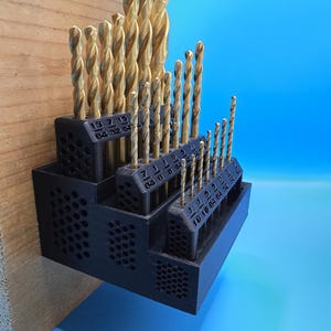 May include: A black, tiered drill bit organizer with multiple gold-colored drill bits. The organizer is mounted on a wooden surface and has labels with fractional measurements. The background is a gradient of blue.