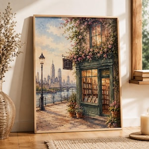 May include: A framed wall art print depicting a bookshop with a green storefront, overflowing with pink flowers, situated along a riverbank. In the background, a city skyline with skyscrapers is visible across the water under a soft sky.