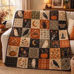 May include: A quilted throw blanket featuring a patchwork grid of autumn-themed illustrations including owls, black cats, crows, pumpkins, crescent moons, mushrooms, and floral motifs in a colour palette of orange, brown, black, and cream.