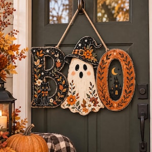 May include: A decorative hanging sign spelling BOO, featuring a black letter B with a cat, a white ghost wearing a witch hat in the centre, and an orange letter O with a haunted house silhouette, all suspended by a thick jute rope cord.