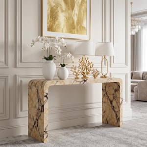Könnte beinhalten: Eine Konsole in Marmoroptik mit geschwungenem Design, verziert mit weißen Vasen mit Orchideen, goldenen Korallen-Skulpturen und zwei Lampen mit weißen Schirmen. Ein goldgerahmtes abstraktes Kunstwerk hängt darüber, wodurch ein elegantes Interieur entsteht.