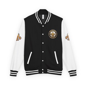 May include: A black varsity jacket with white sleeves, featuring a circular New Orleans Saints logo on the left chest and gold fleur-de-lis emblems on both sleeves. The jacket has a snap-button front, striped ribbed collar, and cuffs.