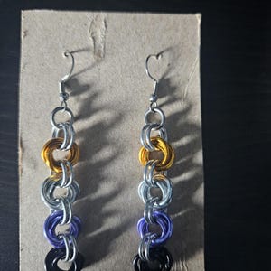 Könnte beinhalten: Ein Paar Hängeohrringe im Kettengeflecht-Design mit ineinandergreifenden Metallringen. Die Ringe sind in einem vertikalen Muster mit abwechselnden Farben in Gelb, Silber, Violett und Schwarz angeordnet und mit silberfarbenen Ohrhaken versehen.