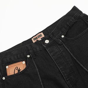 May include: Black denim jeans featuring a tan leather patch on the waistband with the text DENIM GENES and a smaller tan leather patch on the front pocket with a black letter C and a star. The jeans include silver metal rivets and white stitching.
