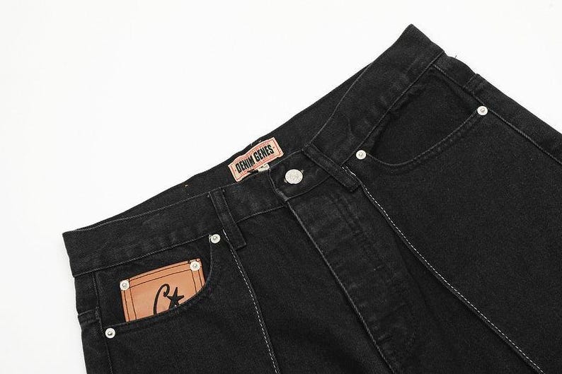 May include: Black denim jeans featuring a tan leather patch on the waistband with the text DENIM GENES and a smaller tan leather patch on the front pocket with a black letter C and a star. The jeans include silver metal rivets and white stitching.