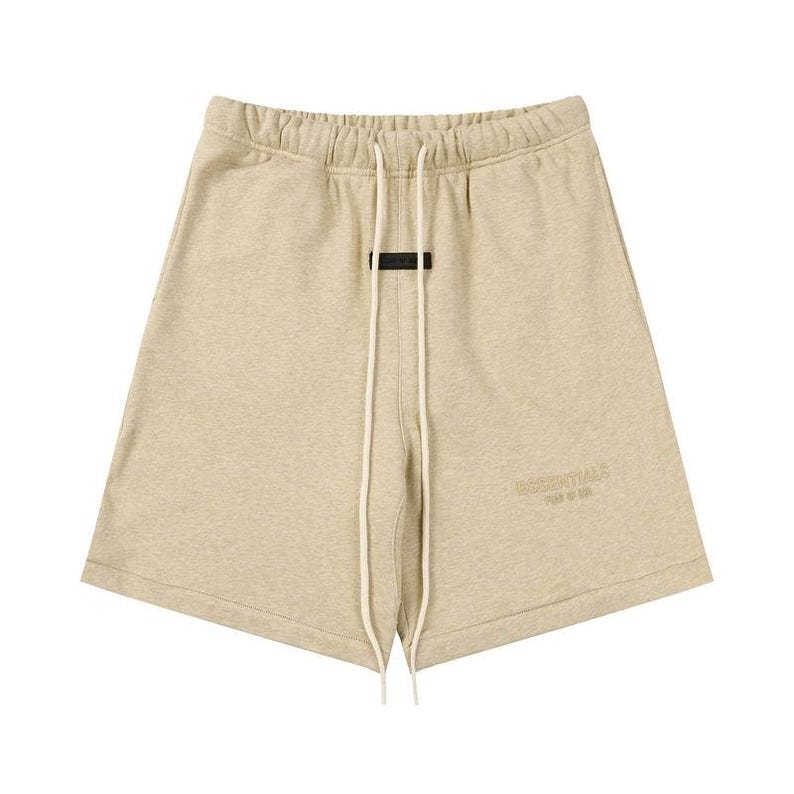 May include: Beige sweat shorts featuring an elastic waistband with long off-white drawstrings. A small black rectangular patch is centered below the waistband, and the word ESSENTIALS FEAR OF GOD is embossed on the lower left leg area.