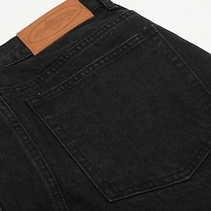 May include: Black denim jeans featuring a large, rectangular back pocket and a tan leather patch sewn onto the waistband. The patch displays an embossed oval design with text and a graphic illustration of a submarine, with visible stitching.