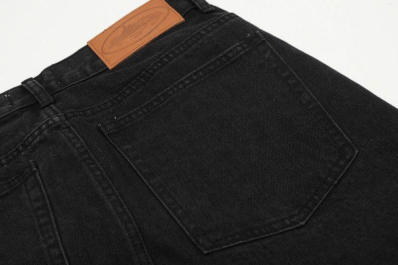 May include: Black denim jeans featuring a large, rectangular back pocket and a tan leather patch sewn onto the waistband. The patch displays an embossed oval design with text and a graphic illustration of a submarine, with visible stitching.