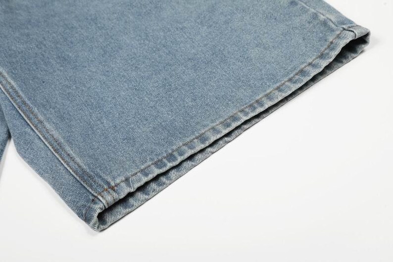 Peut inclure: Un gros plan sur l'ourlet d'un jean en denim bleu clair. Le tissu pr&eacute;sente une texture en serg&eacute; classique avec des coutures visibles le long du bord inf&eacute;rieur, mettant en valeur la coupe droite et la construction durable du mat&eacute;riau en denim.