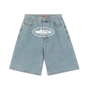 May include: Light blue denim shorts featuring a high-waisted design with a button fly closure. A white oval graphic with a landscape illustration is centered on the front. A small white embroidered logo is on the coin pocket and a label reads DENIM GENES.