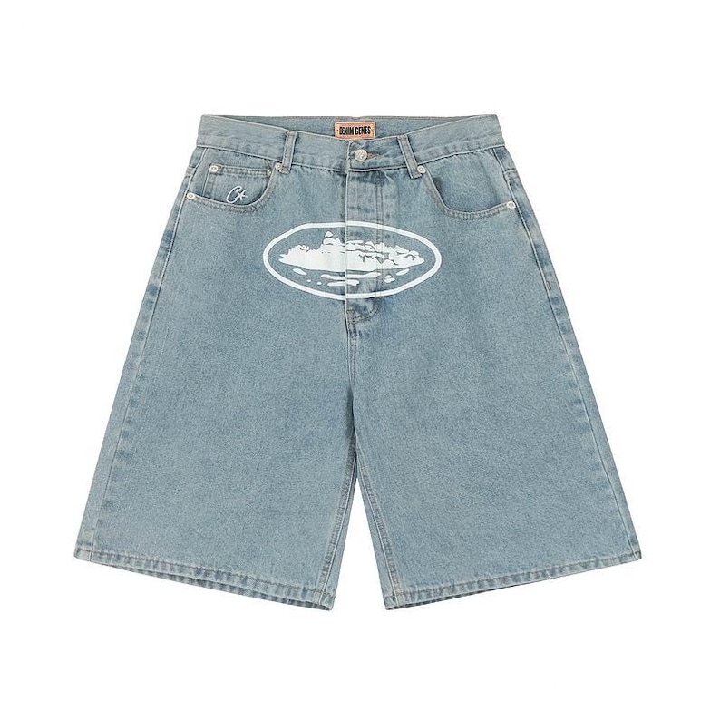 May include: Light blue denim shorts featuring a high-waisted design with a button fly closure. A white oval graphic with a landscape illustration is centered on the front. A small white embroidered logo is on the coin pocket and a label reads DENIM GENES.