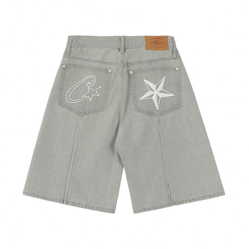 May include: Light gray denim shorts featuring a high-waisted design with a tan leather patch on the back waistband. The back pockets display white embroidered details, including a stylized letter C with a star on the left and a large star on the right.