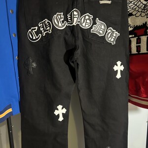May include: Black denim jeans with white cross and "CHROME" patches. The jeans have a classic five-pocket design and a leather patch on the back. The patches are embroidered with a gothic font. The jeans are a dark wash.