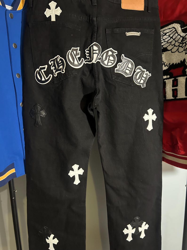 May include: Black denim jeans with white cross and "CHROME" patches. The jeans have a classic five-pocket design and a leather patch on the back. The patches are embroidered with a gothic font. The jeans are a dark wash.