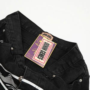 May include: Black denim jeans featuring a waistband with belt loops and silver-tone metal rivets. A rectangular purple and yellow tag is attached, displaying the text DENIM GENES, LONDON, ENGLAND, 2016, and additional decorative graphic elements.