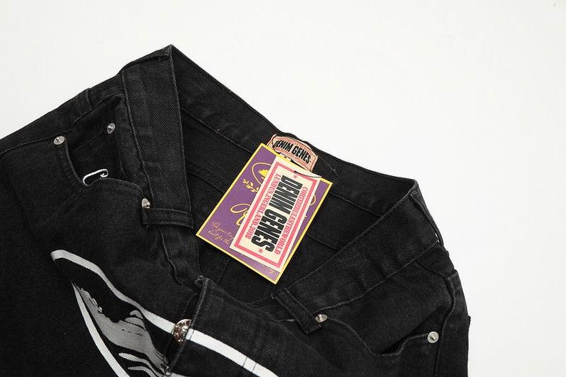 May include: Black denim jeans featuring a waistband with belt loops and silver-tone metal rivets. A rectangular purple and yellow tag is attached, displaying the text DENIM GENES, LONDON, ENGLAND, 2016, and additional decorative graphic elements.