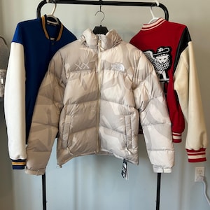May include: A display of outerwear, including a light beige puffer jacket with a camouflage pattern and the North Face logo, and two varsity jackets. One varsity jacket is blue with white sleeves, and the other is red with black sleeves.