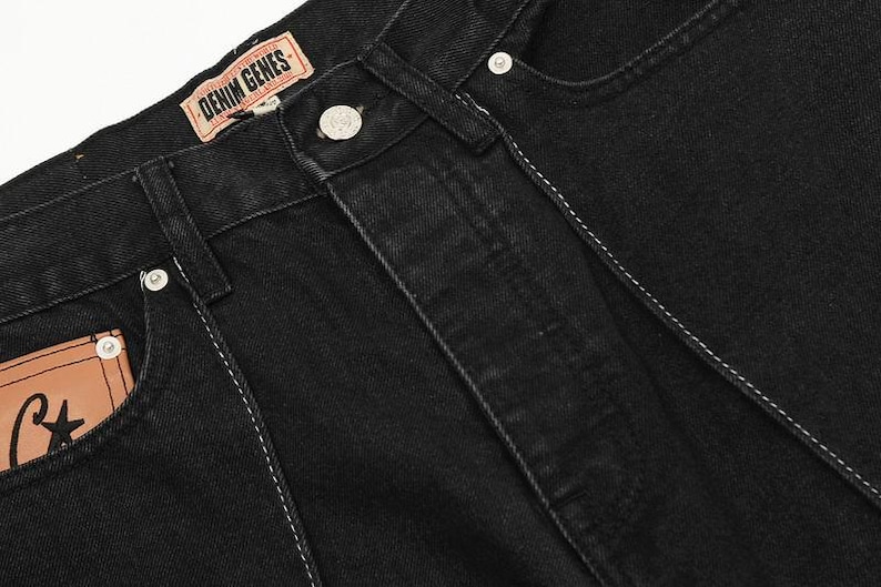 May include: Black denim jeans featuring a tan leather brand patch on the waistband with the text DENIM GENES, a silver metal button closure, and a tan leather pocket accent with a black star and letter C graphic, accented by white stitching.