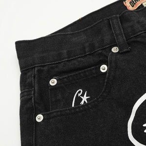 May include: Black denim jeans featuring a high-waisted design with a waistband, belt loops, and silver-toned metal rivets. A small white embroidered star logo is visible on the coin pocket, and a large white graphic print is on the front.