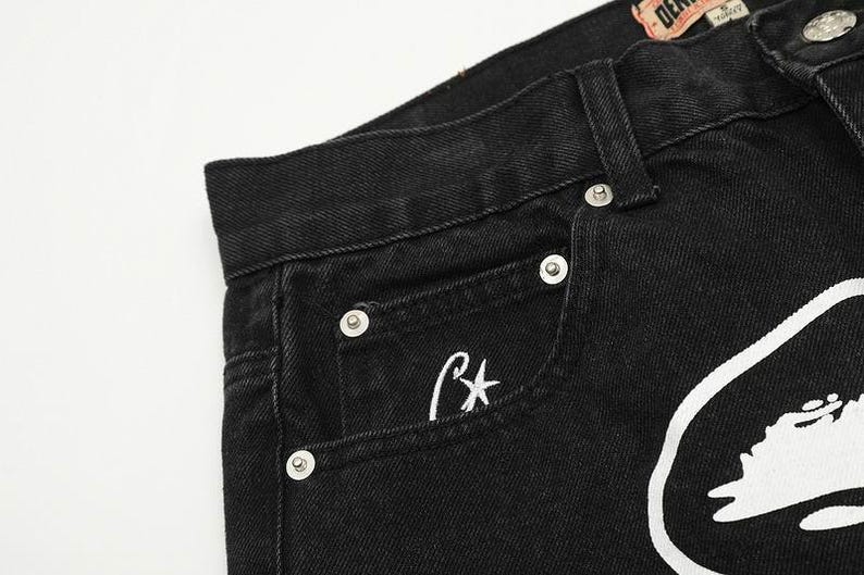 May include: Black denim jeans featuring a high-waisted design with a waistband, belt loops, and silver-toned metal rivets. A small white embroidered star logo is visible on the coin pocket, and a large white graphic print is on the front.