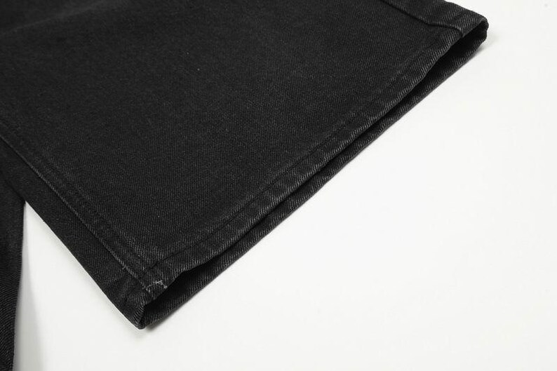 May include: A close-up view of the hem of a black denim garment. The fabric features a visible twill weave texture with sturdy, tonal stitching along the folded edge. The material appears to be a durable cotton denim with a matte finish.