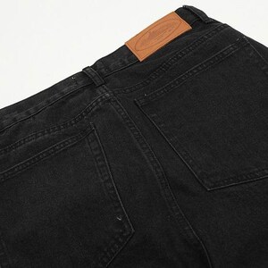 May include: Black denim jeans featuring two back pockets, belt loops, and a tan leather patch on the waistband. The patch displays an embossed oval logo with a central graphic design. The fabric has a visible twill texture and tonal stitching.