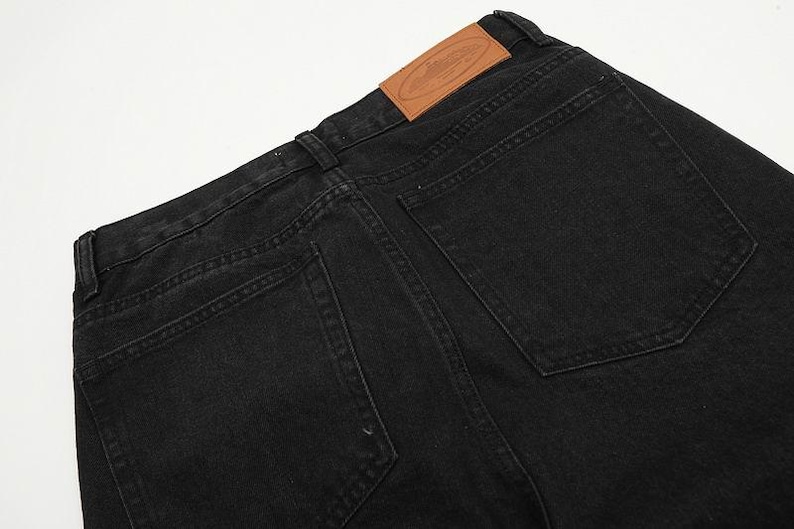 May include: Black denim jeans featuring two back pockets, belt loops, and a tan leather patch on the waistband. The patch displays an embossed oval logo with a central graphic design. The fabric has a visible twill texture and tonal stitching.
